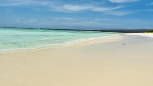 Une plage à l’eau turquoise et limpide, bordée d’une vaste étendue de sable fin, sous un ciel partiellement nuageux.