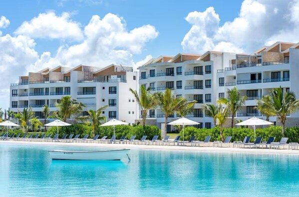Un hôtel moderne doté de nombreux balcons, d’un espace piscine avec des chaises longues et des parasols, et surplombant un ciel d’un bleu éclatant.