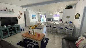 Uma sala de estar com televisão, uma mesa de centro com um vaso de flores e um sofá. Há também um espaço de jantar com mesa e cadeiras, e uma cozinha ao fundo.