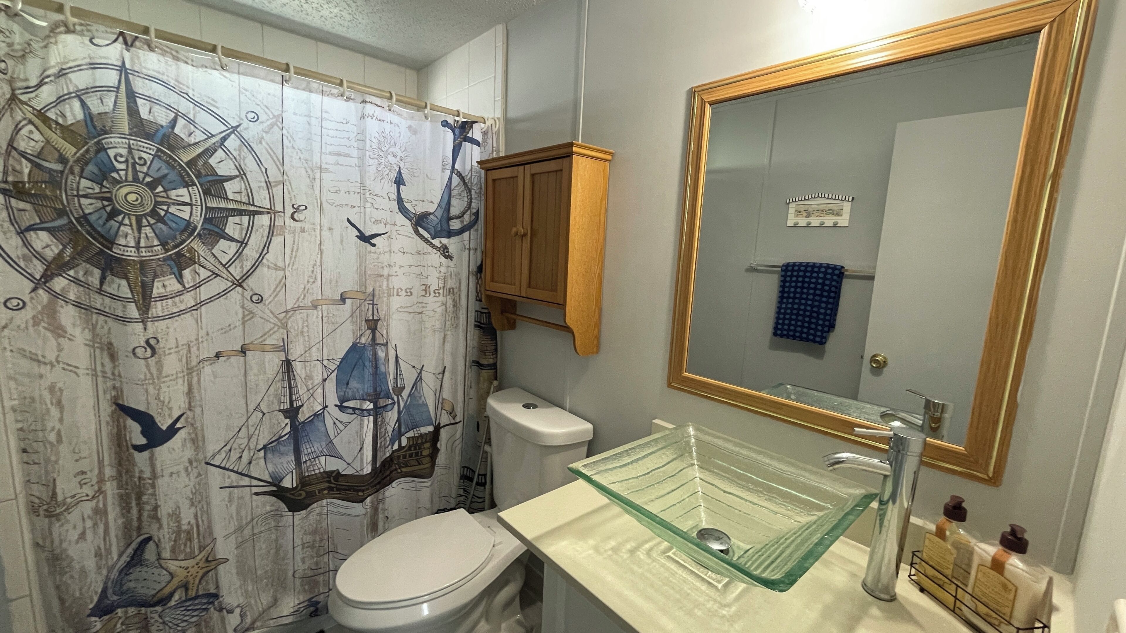 A bathroom with a nautical-themed shower curtain, a glass sink, a wooden cabinet, and a mirror.