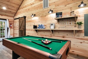 A pool table with cues and balls, a fire extinguisher, and a shelf with glasses and a book in a wooden room.