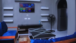 A modern living room with a flat-screen TV mounted on the wall, a black fan, and a blue sofa. - Founders Apartments (Kabwe)