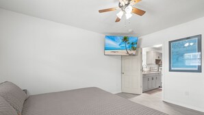 A bedroom with a bed, a ceiling fan, a TV on the wall, and a kitchen in the background. - Walk To Beach, North End Restaurants, Night Life Steps Away! (Fort Myers Beach)