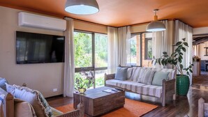 A modern living room with a sofa, coffee table, and a TV. There are large windows with curtains, a plant, and a view of greenery outside. - Tuscan Farmhouse (Don Valley)