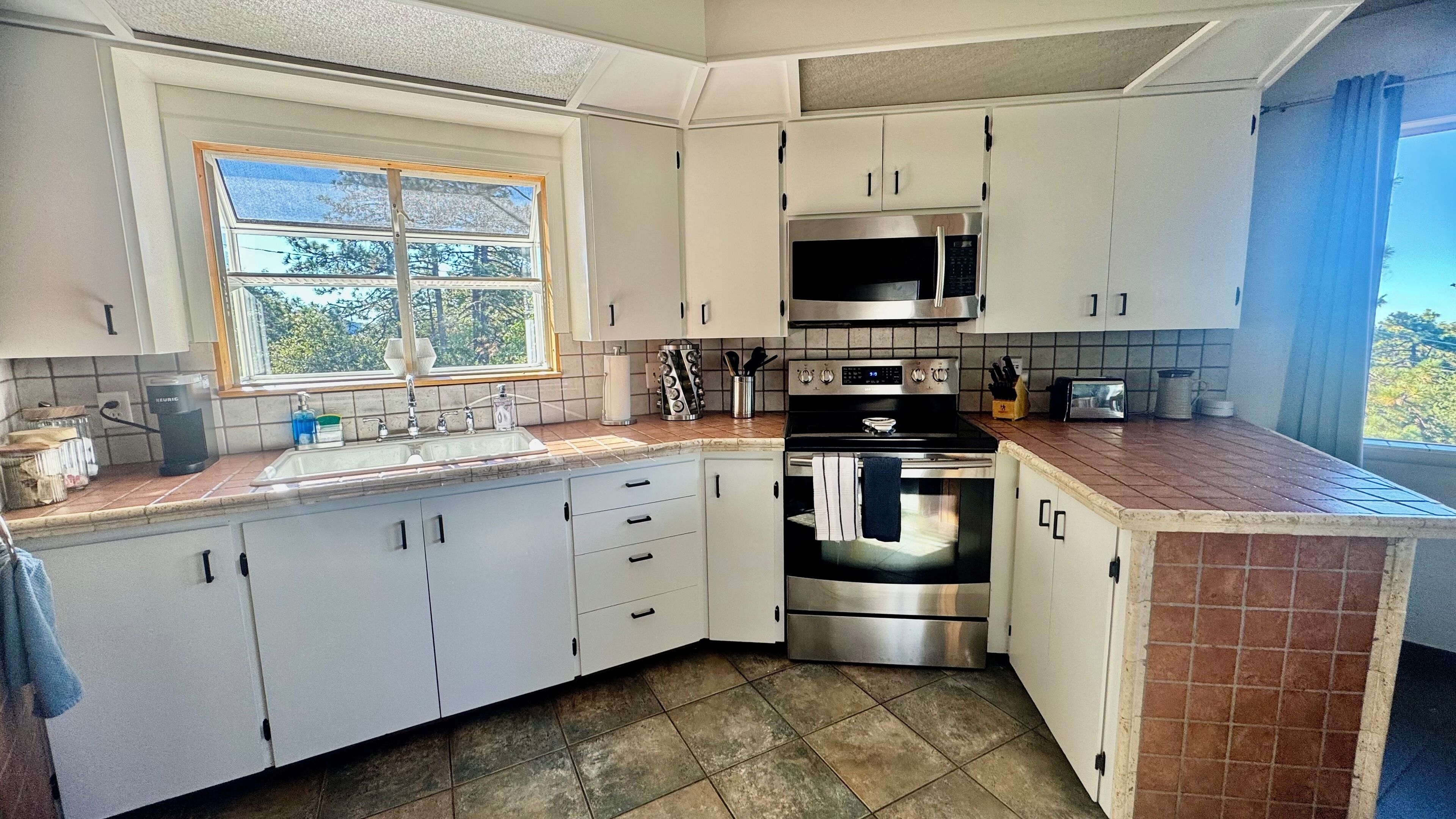 A kitchen with white cabinets, a stainless steel oven, a microwave, and a window with a view of trees.