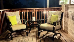 An outdoor deck with two chairs and a table, surrounded by wooden railings and a view of trees.