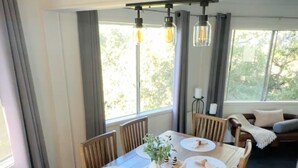 A dining area with a wooden table set for four, surrounded by chairs, with a view of greenery outside.