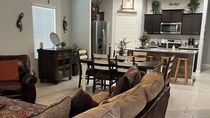 A modern living room with a large sectional sofa, a dining area with wooden chairs, and a kitchen with dark cabinets and stainless steel appliances. - Saguaro Bloom Beauty (Marana)