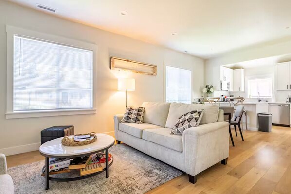 A modern living room with a sofa, coffee table, and a small side table. There is a kitchen area with white cabinets and a dining table. - Downtown Charm Meets Waterfront Walk to Everything (Vancouver)