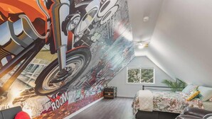 A bedroom with a sloped ceiling, a mural of a motorcycle, a bed with floral bedding, a red heart-shaped pillow, and a small table.