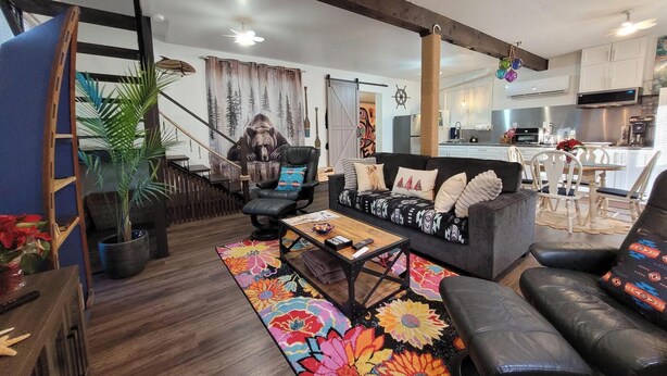 A modern living room with a sofa, armchairs, a coffee table, and a colorful rug. A staircase with a wooden railing leads to an upper level.