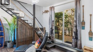 A modern living room with a staircase, a large window with curtains, a lounge chair, and a potted plant.