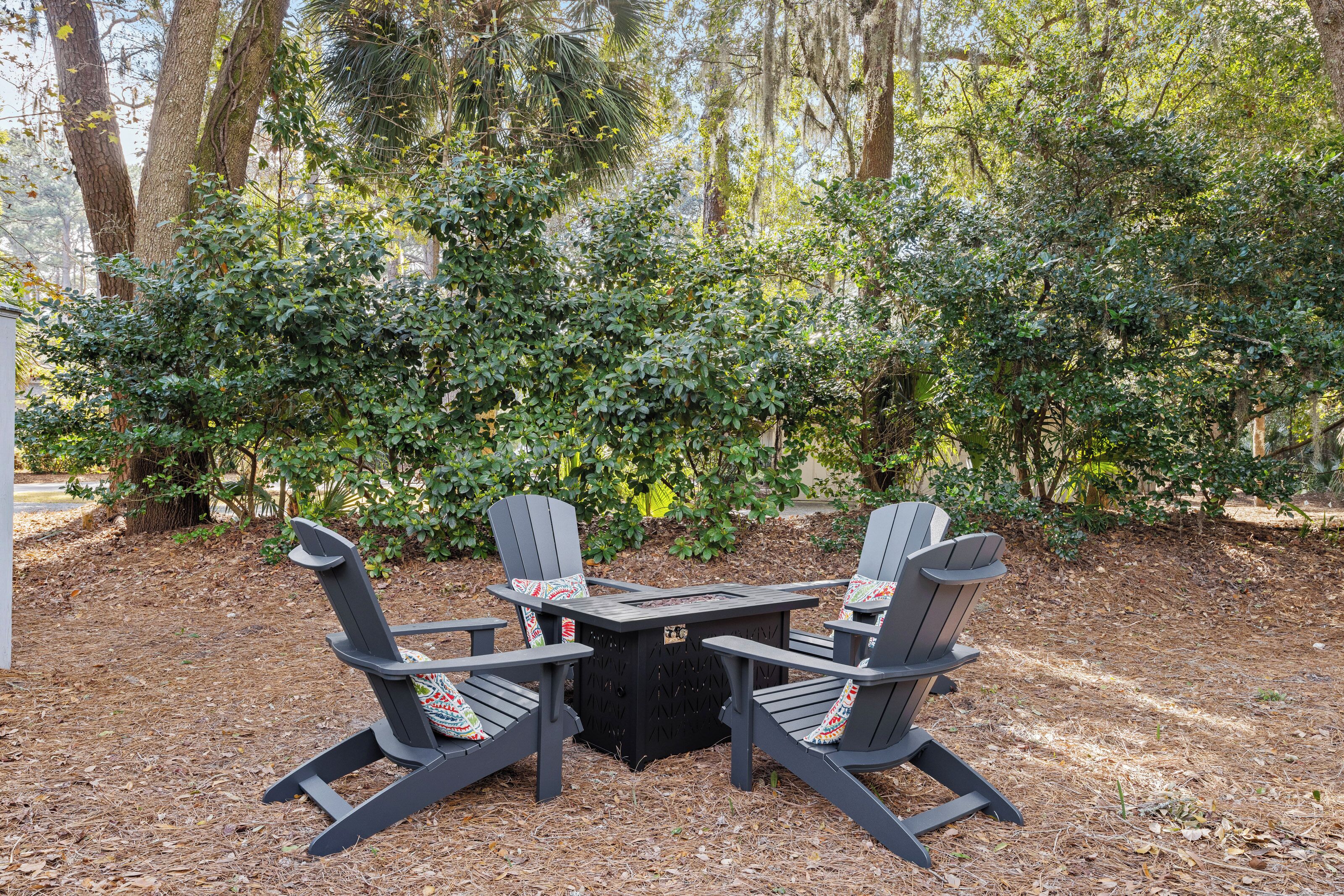 Adirondack-Stühle um einen quadratischen Tisch in einem Gartenbereich, im Hintergrund üppiges Grün.