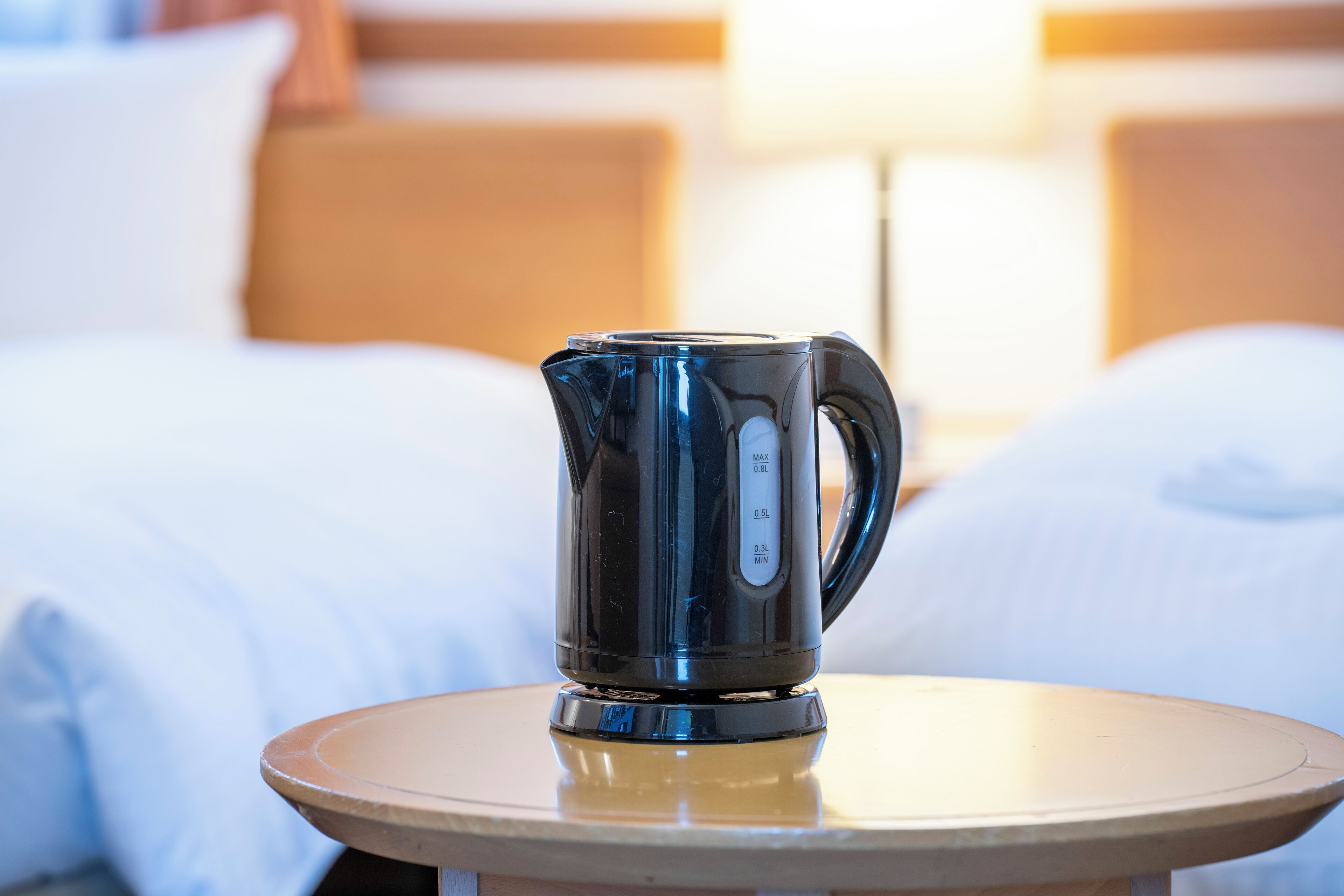 A black electric kettle on a round table in a hotel room with a bed and a nightstand in the background.