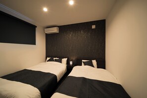 Habitación de hotel con dos camas, una pared oscura con textura, un equipo de aire acondicionado y una mesita con una lámpara.