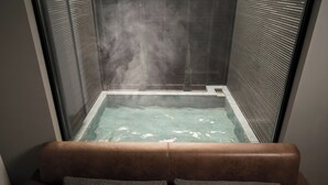 A steam-filled spa room with a glass-enclosed hot tub and a brown leather sofa with gray cushions.