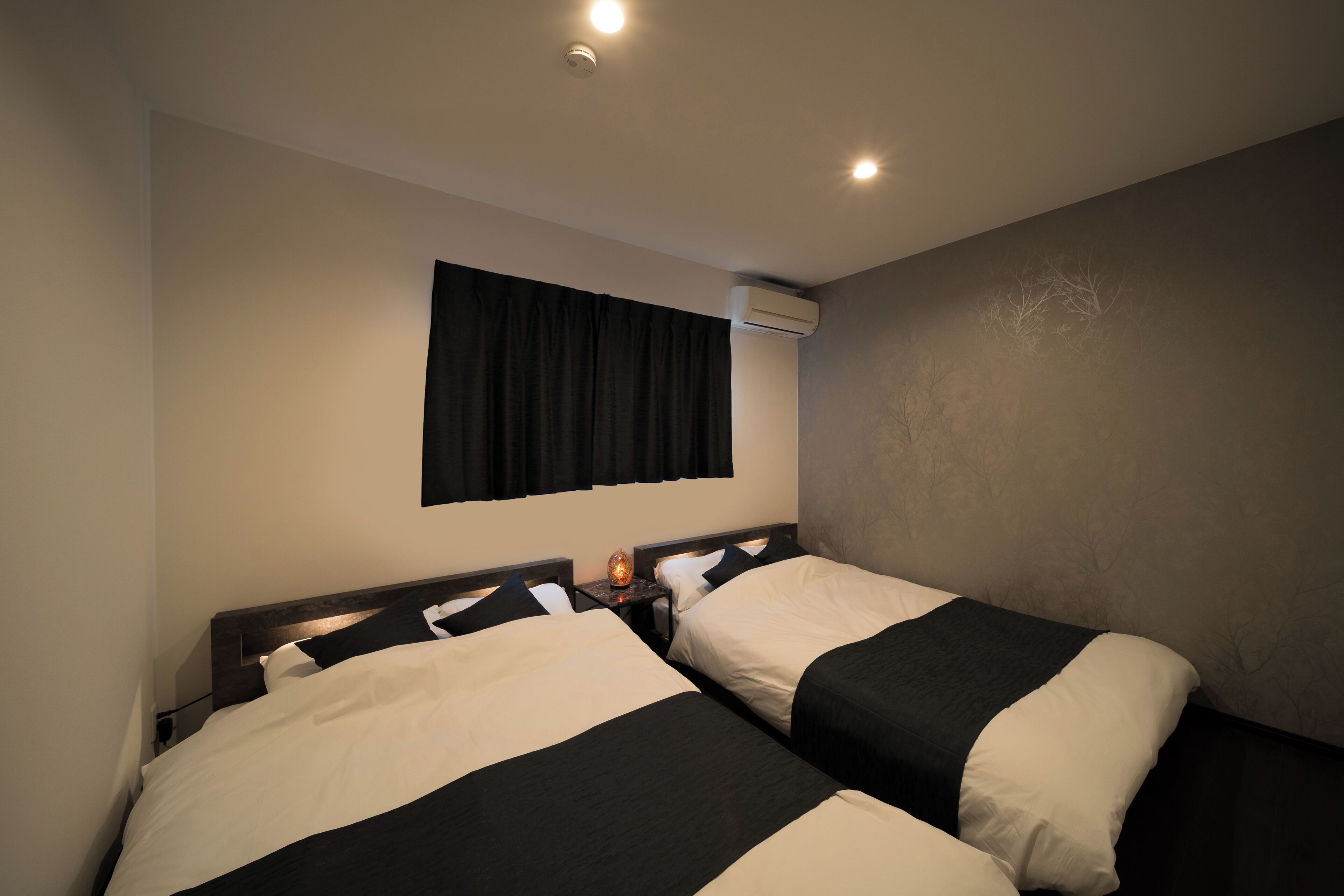 A hotel room with two beds, a nightstand, an air conditioning unit, and a window with curtains.