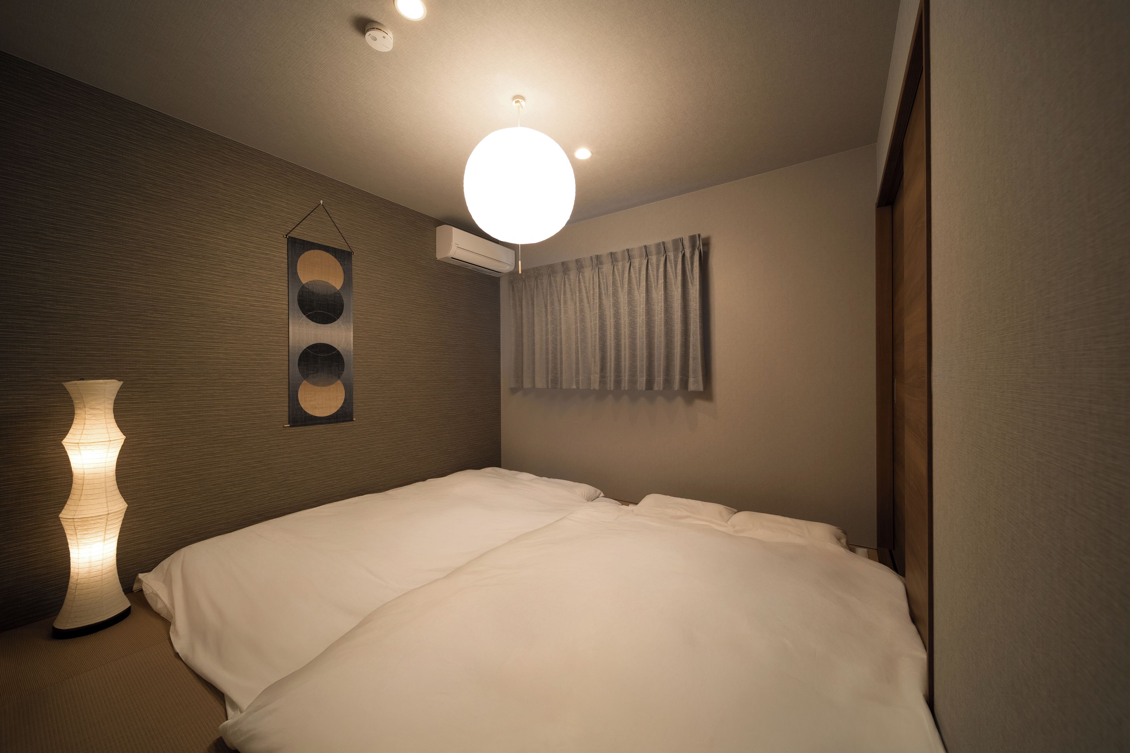 A neatly made bed in a hotel room with a wall-mounted air conditioner, a hanging light fixture, and a curtain on the window.