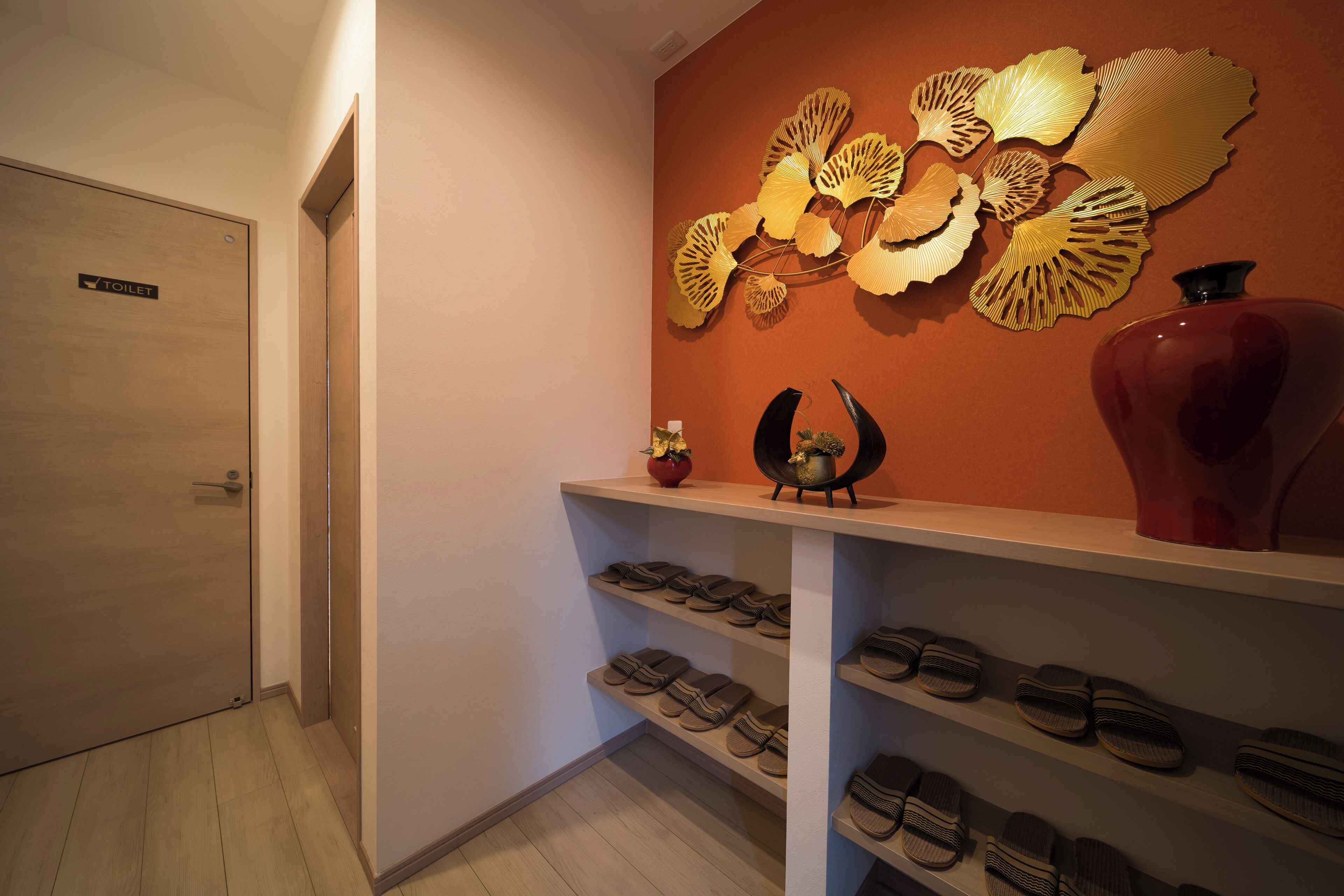 A hallway with a wooden door labeled "TOILET," a wall-mounted decorative piece, a shelf with shoes, and a vase.
