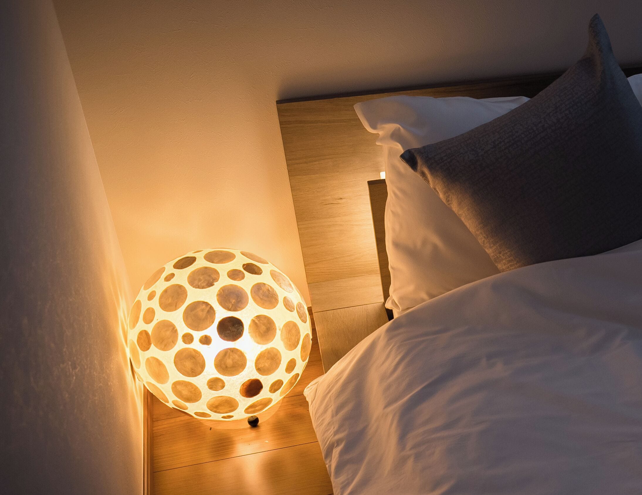 A bedroom with a wooden floor, a white bed with pillows, and a unique circular-patterned lamp.