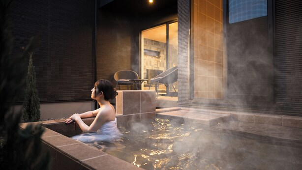 A person relaxing in a hot tub outside a modern building with a glass door and a chair visible in the background.