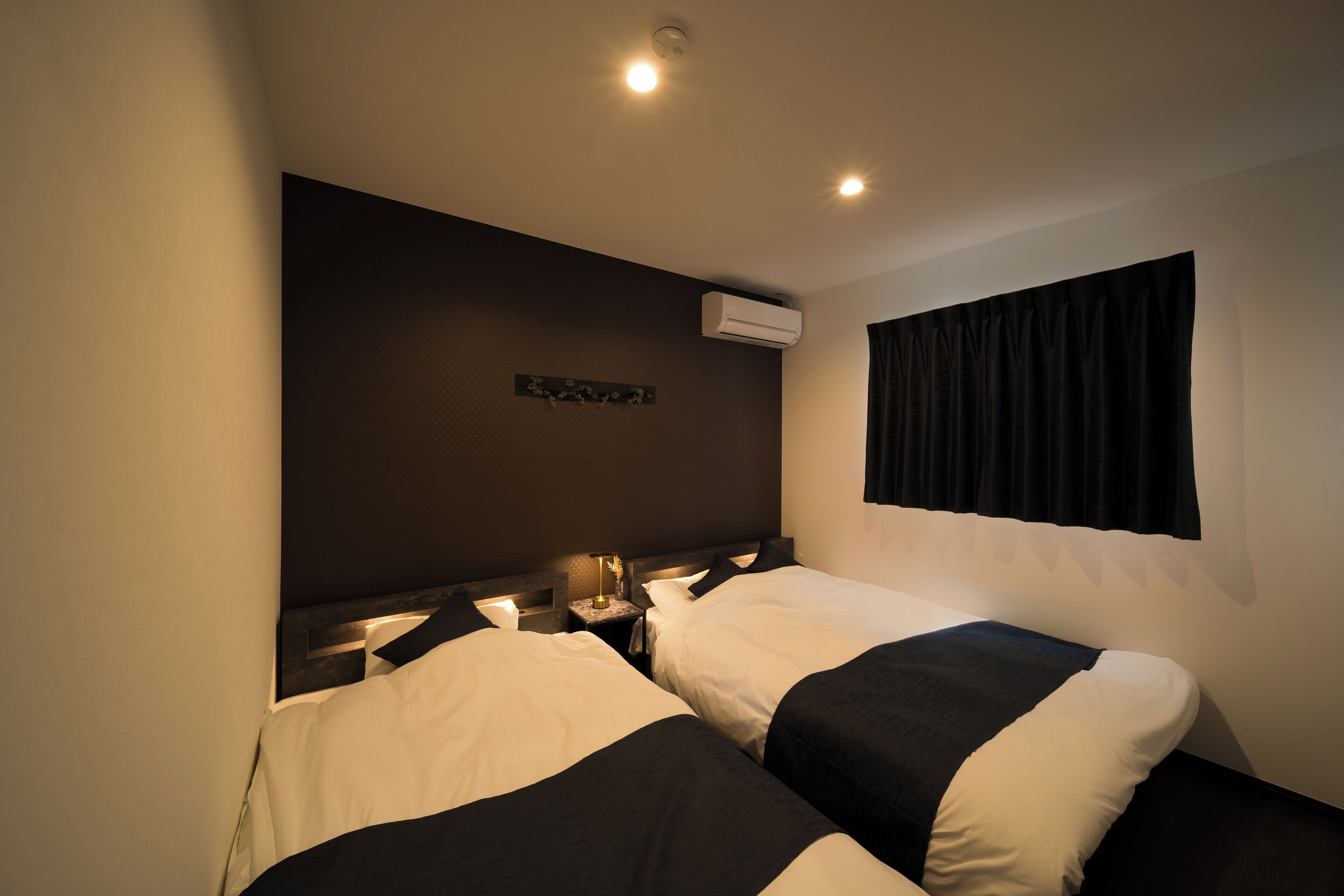 A hotel room with two beds, a nightstand, an air conditioning unit, and a window with curtains.