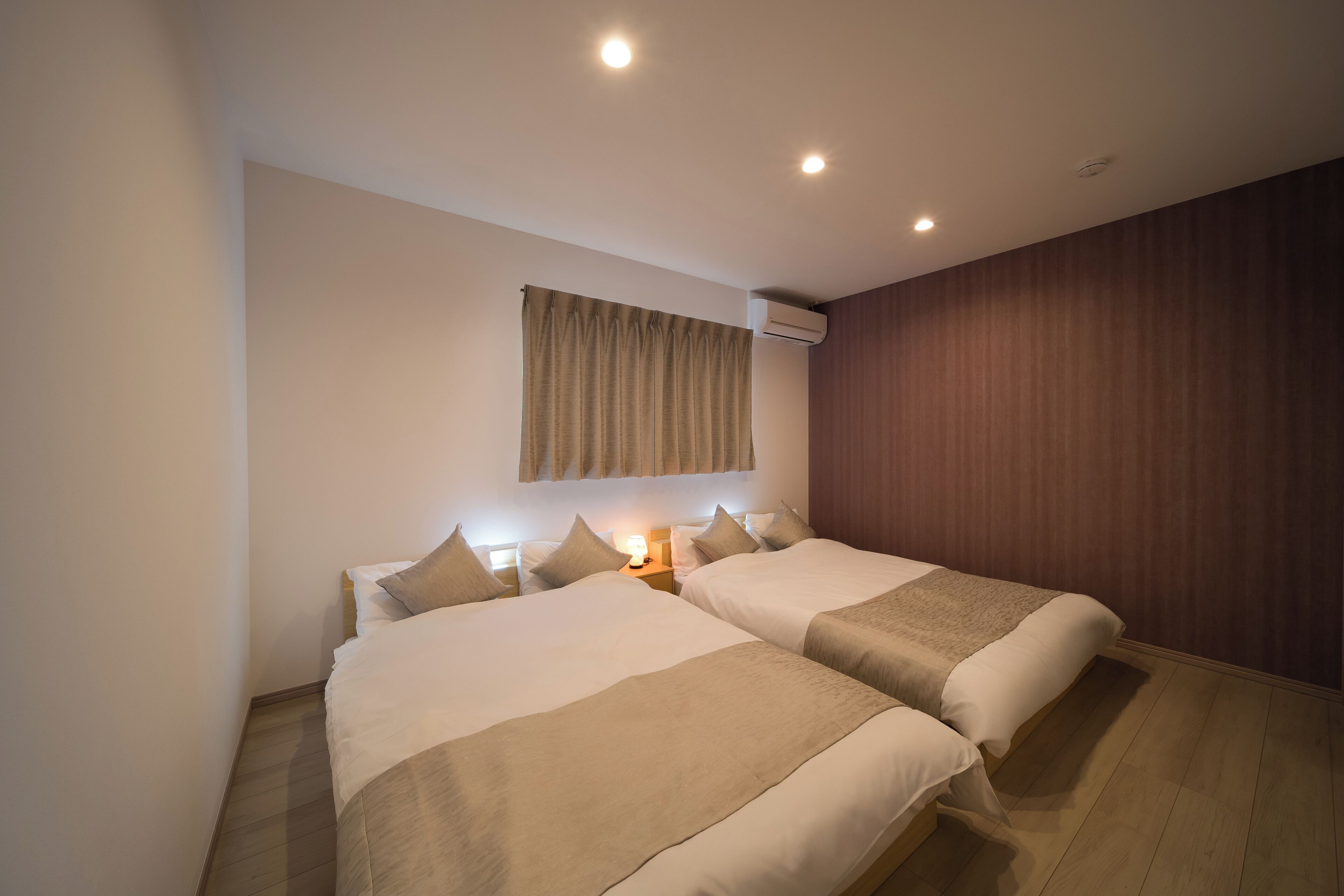 A hotel room with two beds, a wooden floor, a wall-mounted air conditioner, and a window with curtains.
