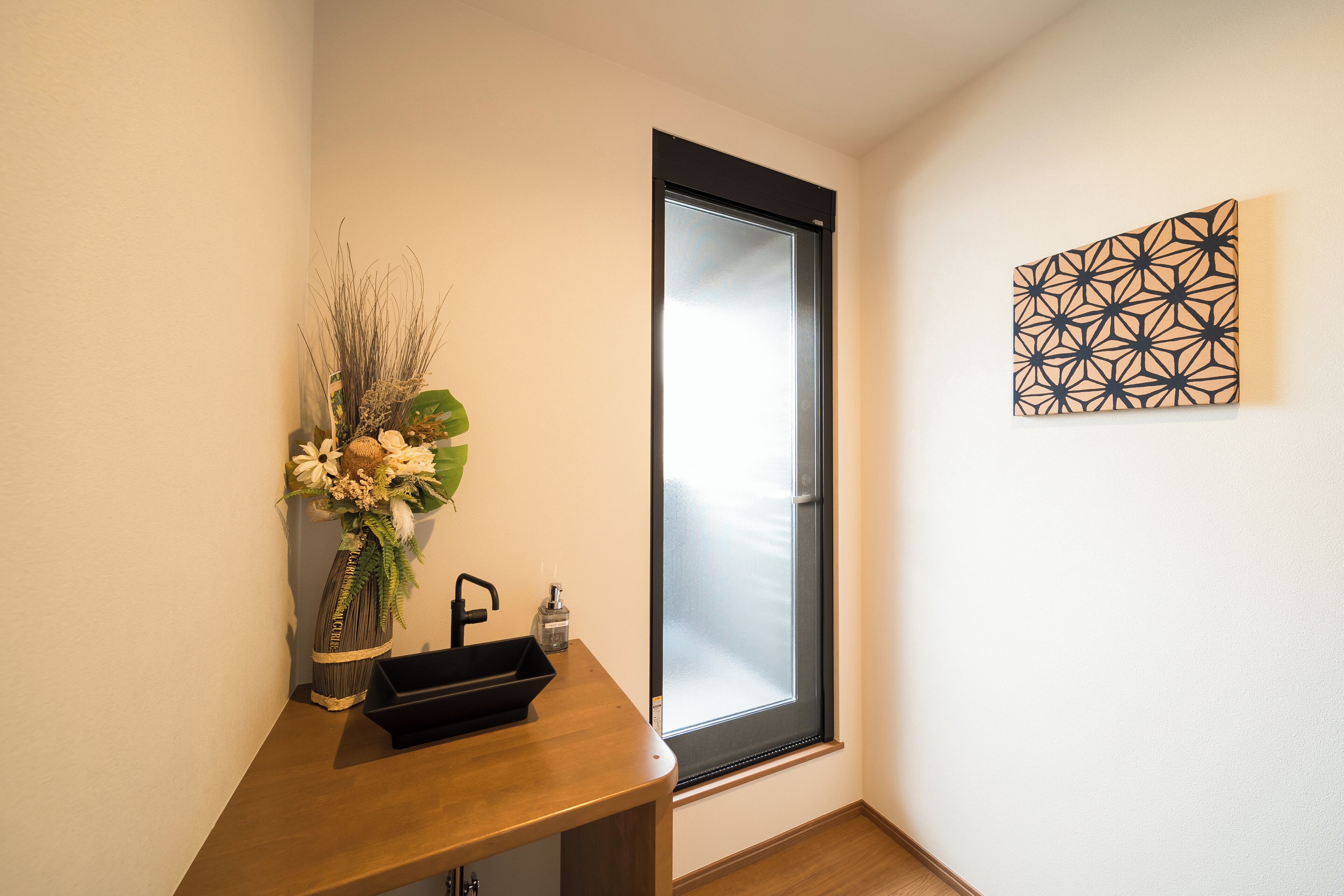 A bathroom with a wooden vanity, a vase with flowers, a black soap dispenser, and a geometric wall art piece.