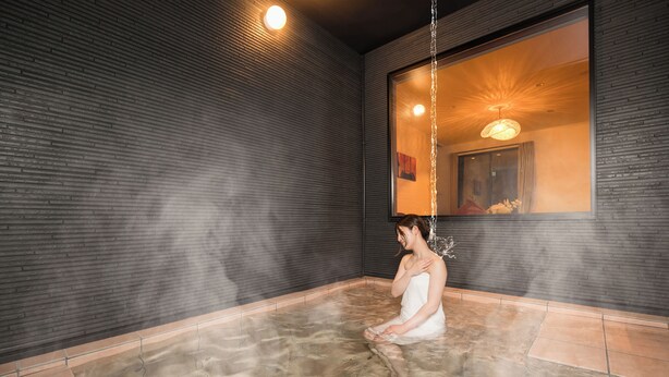 Eine Person sitzt in einer Dampfsauna mit gefliestem Boden und einem großen Spiegel an der Wand.