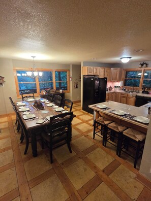 A dining area with a wooden table set for six, a kitchen with wooden cabinets, and a large window with a view of trees.