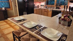 A kitchen with wooden cabinets, a black refrigerator, a dining table set with plates, glasses, and cutlery, and two wicker bar stools.