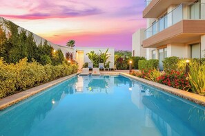Un área de piscina moderna en un hotel, con vista despejada a la piscina, sillones de descanso y follaje circundante.