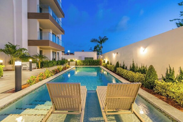 Piscina con sillas de descanso, rodeada de jardines cuidados y un edificio con balcones.