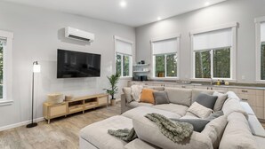 A modern living room with a large flat-screen TV, a beige sectional sofa, a wooden cabinet, and a floor lamp. - House+ADU+GameRoom+Jacuzzi+Pool+Fire pit+Loft (Sisters)