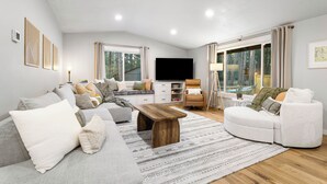 A modern living room with a grey sofa, a wooden coffee table, and a white armchair. There is a flat-screen TV on a stand, a bookshelf, and a window with curtains. - House+ADU+GameRoom+Jacuzzi+Pool+Fire pit+Loft (Sisters)