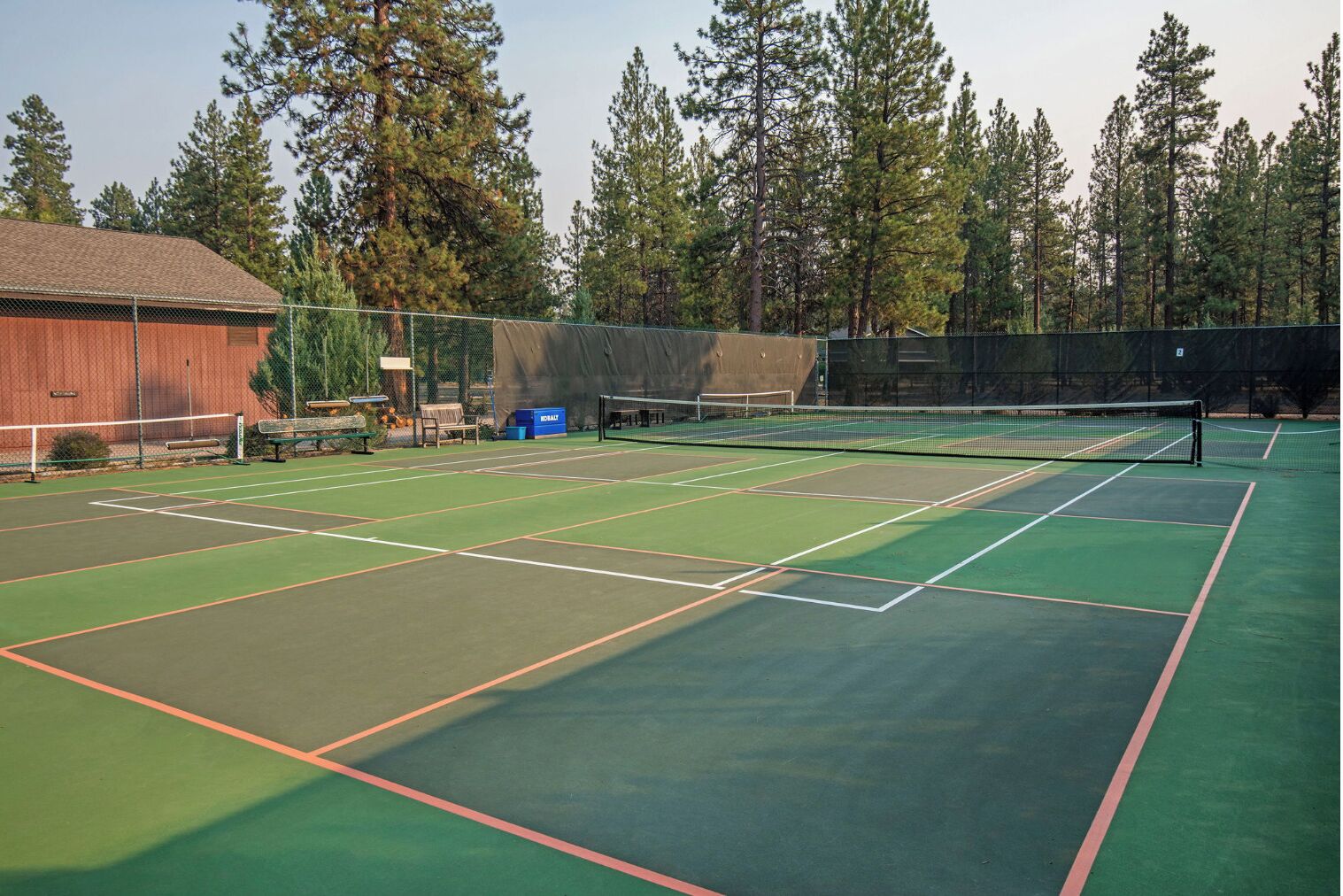 A tennis court with multiple courts, surrounded by trees and a wooden building.