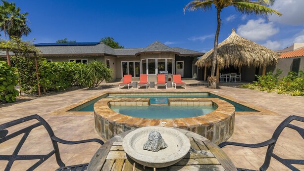 A pool with a hot tub, surrounded by lounge chairs and a dining area with a thatched roof.