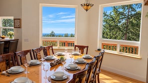 A dining room with a large table set for six, large windows with a view of trees and sky, and a hanging light fixture.