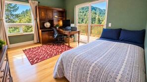 Ein Schlafzimmer mit einem großen Bett, einem Schreibtisch mit Computer, einem Globus und Blick auf einen Wald vom Balkon aus.