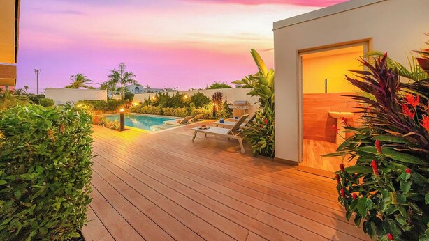 Um deck de madeira com piscina, espreguiçadeiras e um edifício moderno com porta aberta.