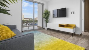 Uma sala de estar moderna com um sofá cinzento, um almofão amarelo, uma janela grande com vista para edifícios, uma televisão de tela plana e um console branco com detalhes amarelos.