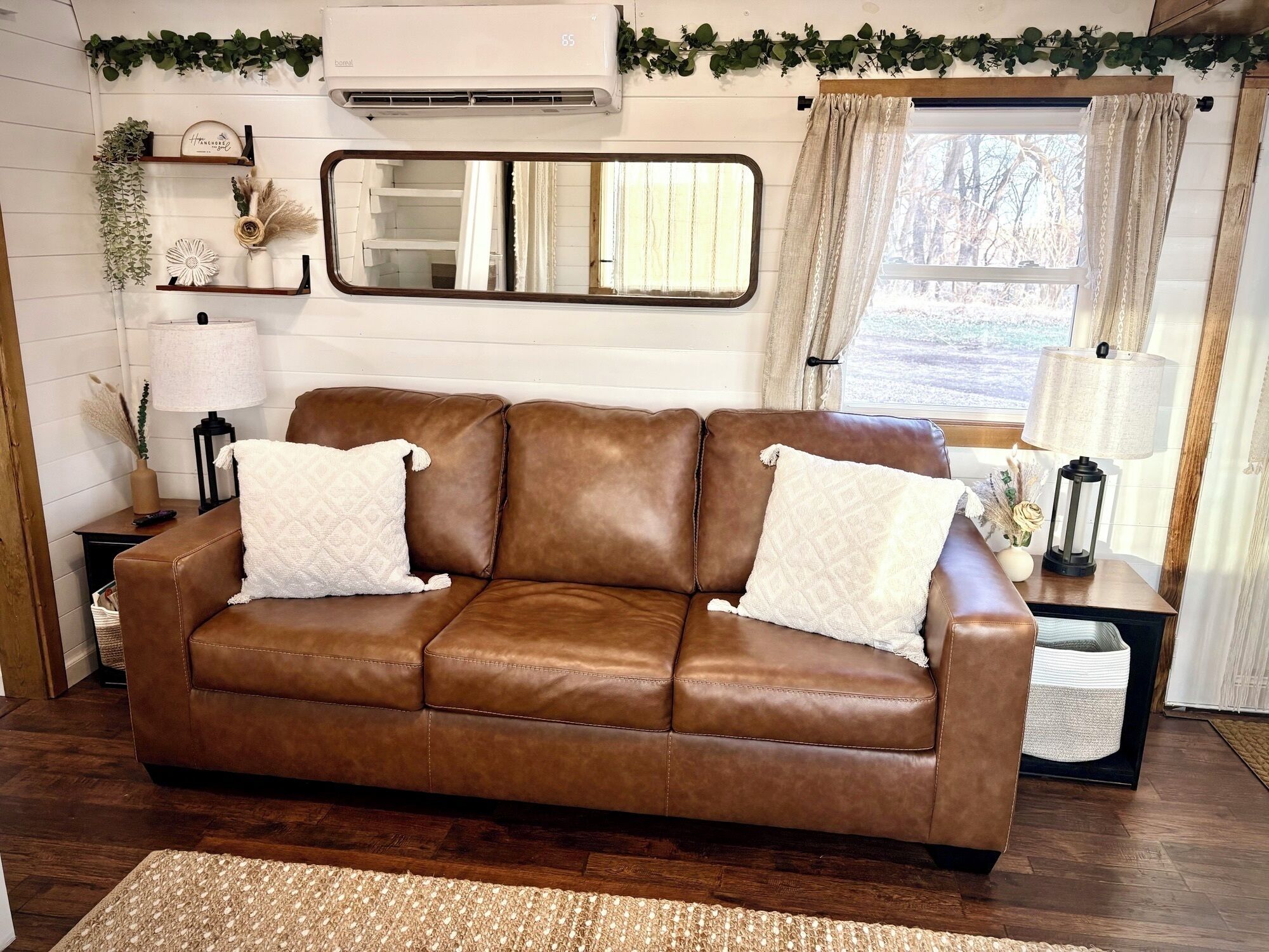 A brown leather sofa with two pillows, a mirror, a lamp, and a small table with decorative items.