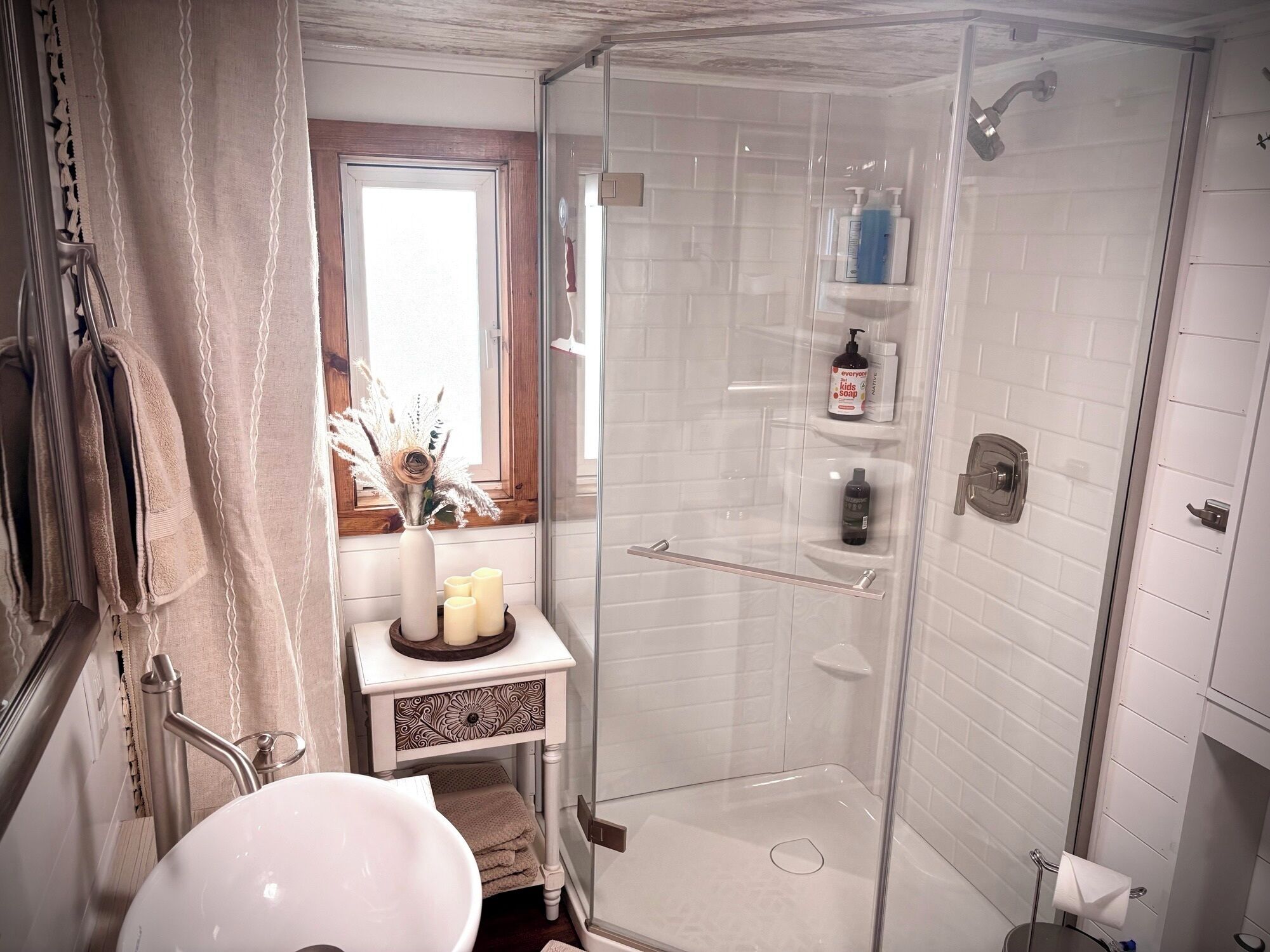 A bathroom with a glass shower enclosure, a white sink, a vanity with a vase and candles, and toiletries on a shelf.