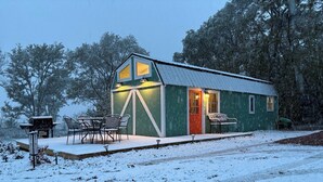 A small, green wooden cabin with a red door, a porch with chairs, and a snow-covered ground. - Secluded Tiny House with Fire Pit (Ponca)