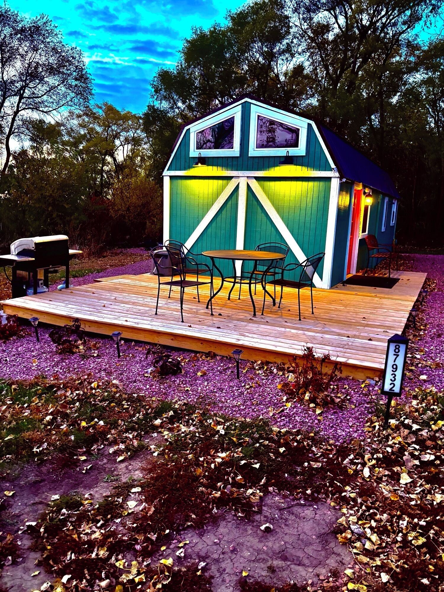Une petite structure en bois aux couleurs vives, avec une table et des chaises, entourée d’une terrasse en bois et d’un jardin où poussent des fleurs violettes.
