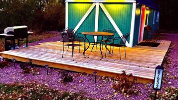 Une petite structure en bois aux couleurs vives, avec une table et des chaises, entourée d’une terrasse en bois et d’un jardin où poussent des fleurs violettes.