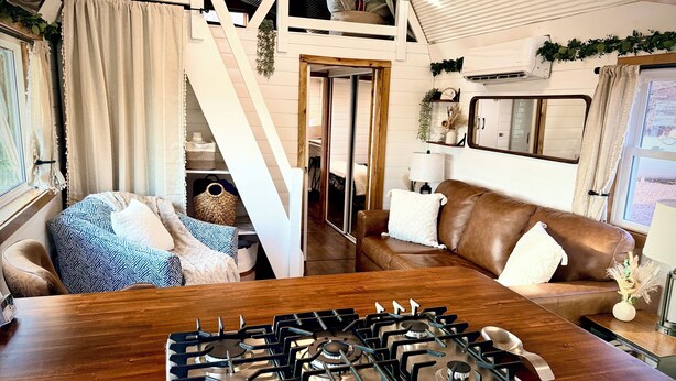 A cozy living area with a brown leather sofa, a wooden dining table, a gas stove, and a staircase leading to an upper level.