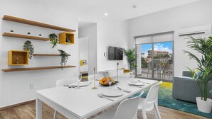 A modern dining area with a white table set for four, surrounded by white chairs. There are wooden shelves with plants on the wall, and a large window offering a view of the city.