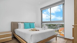 A bedroom with a bed, a nightstand, a window with a city view, and a wooden floor.