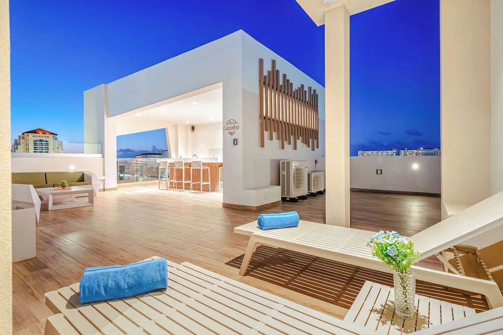 A rooftop lounge area with wooden flooring, white lounge chairs, and a bar area in the background.